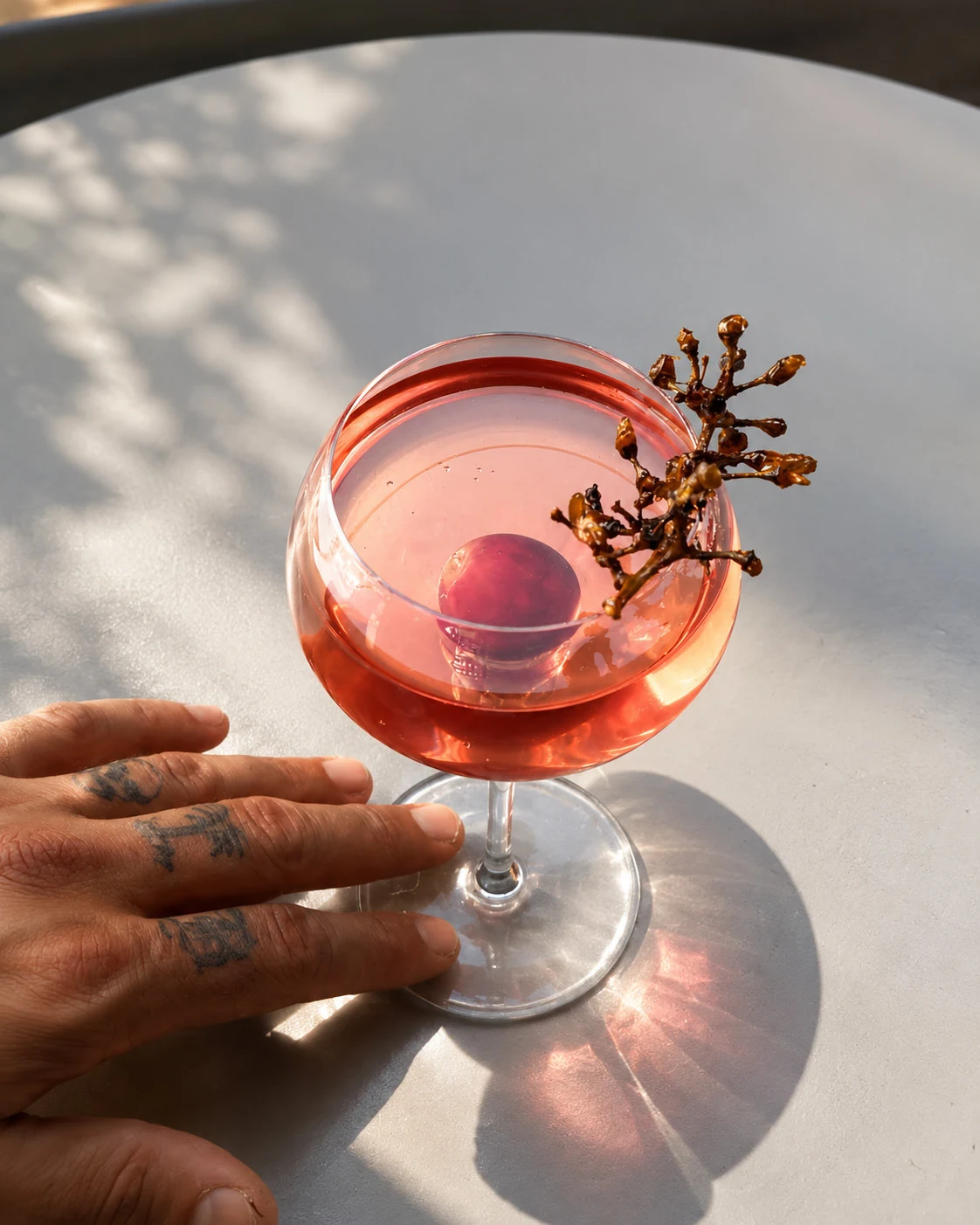 A vivid red cocktail beside a tattooed hand in warm sunlight